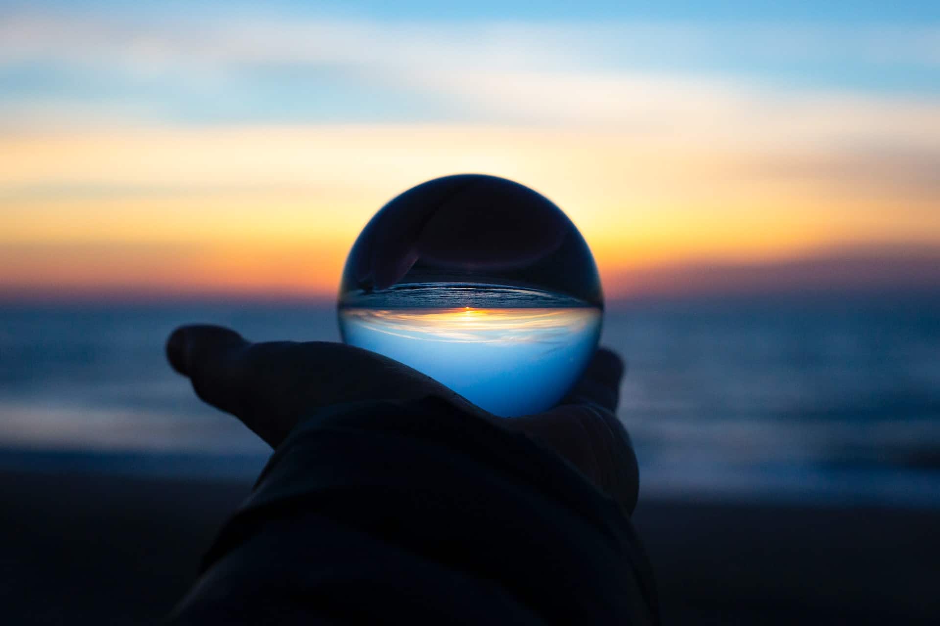 A person holding a glass ball in front of the ocean at sunset A person holding a glass ball in front of the ocean at sunset.